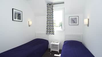 A room with two single beds, a small table with a book, a window with a curtain, and wall-mounted lights.