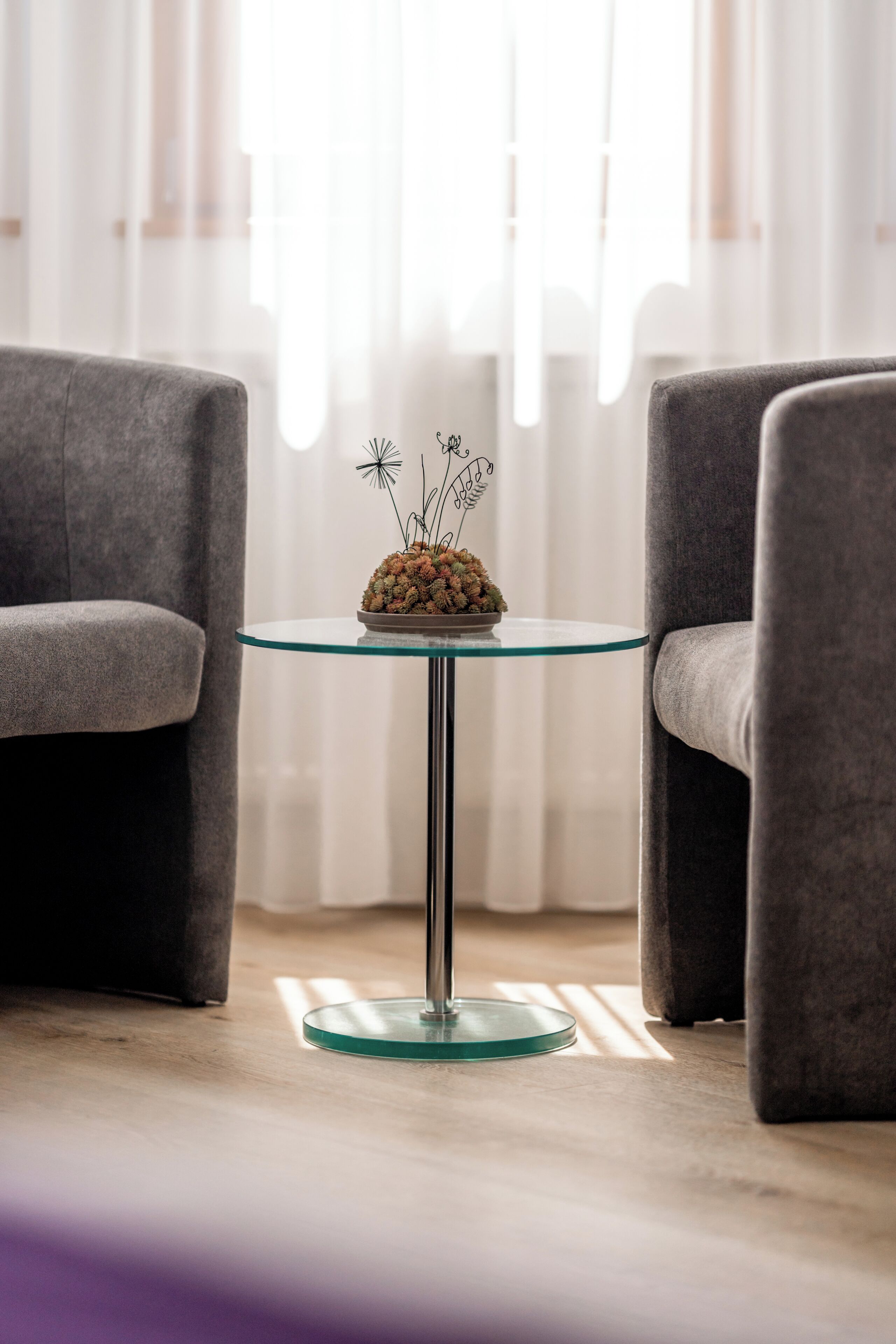 Una habitación moderna con una mesa redonda de cristal en el centro, dos sillones grises y una pequeña planta en maceta sobre la mesa. Cortinas de gasa al fondo.
