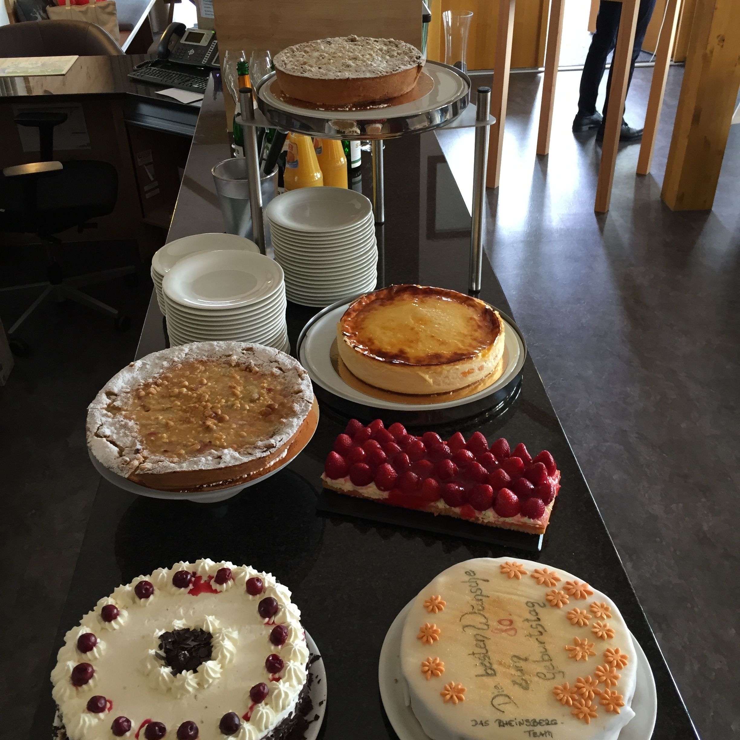 A table with various cakes and pastries, a laptop, and stacked plates.