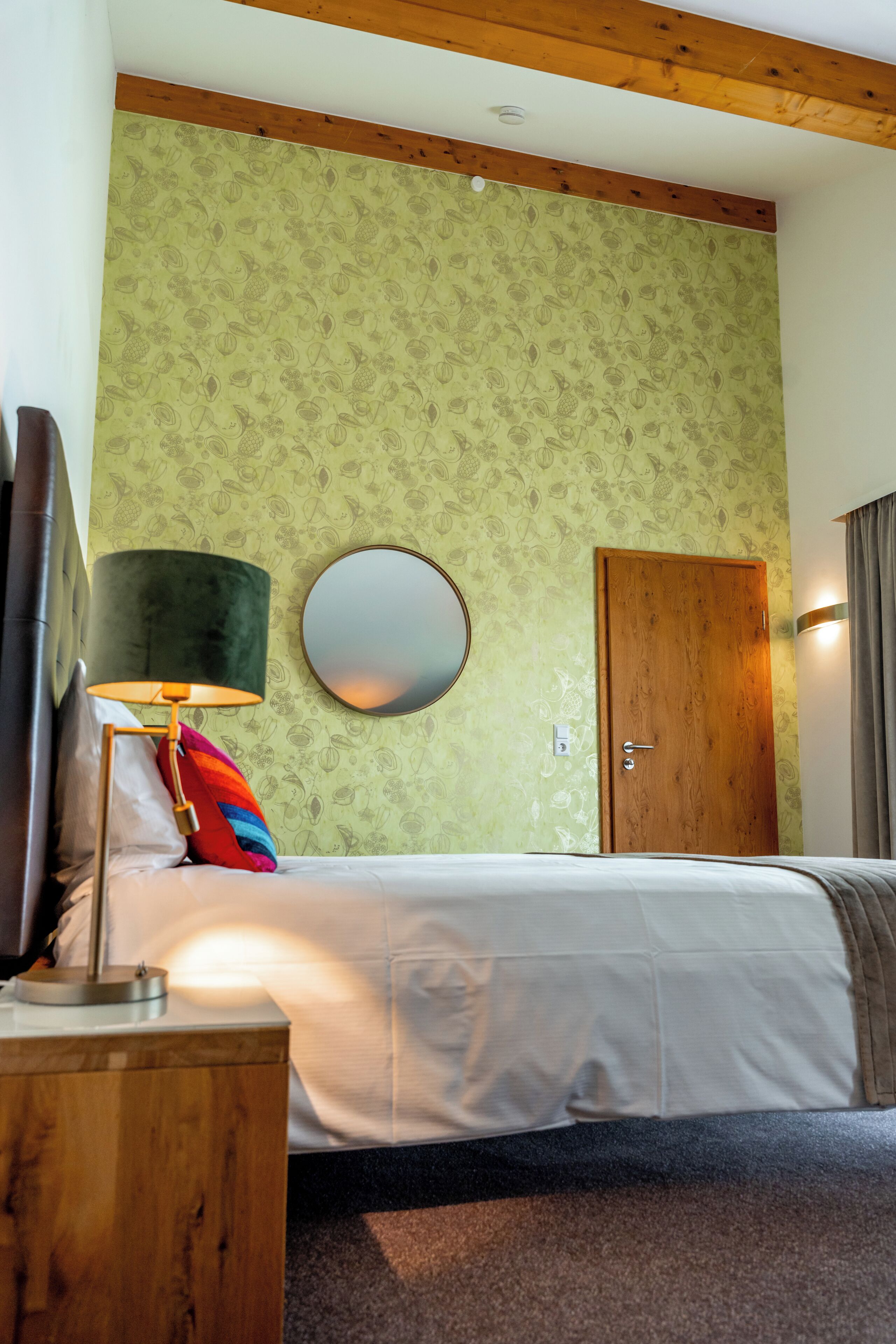 Une chambre d’hôtel avec un lit, un miroir rond, une porte en bois et une lampe.