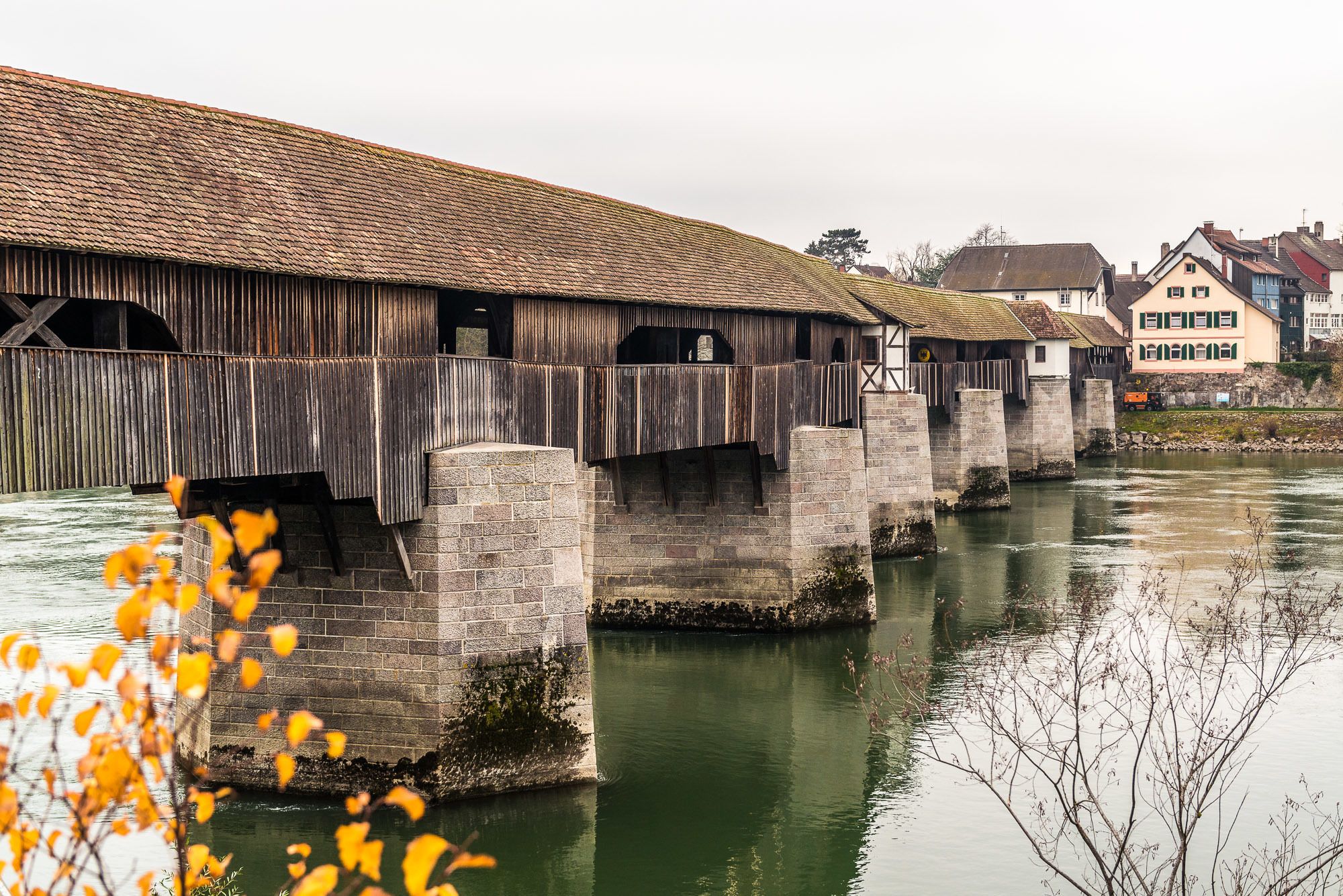 A wooden covered bridge with stone pillars over a calm river, surrounded by traditional buildings.