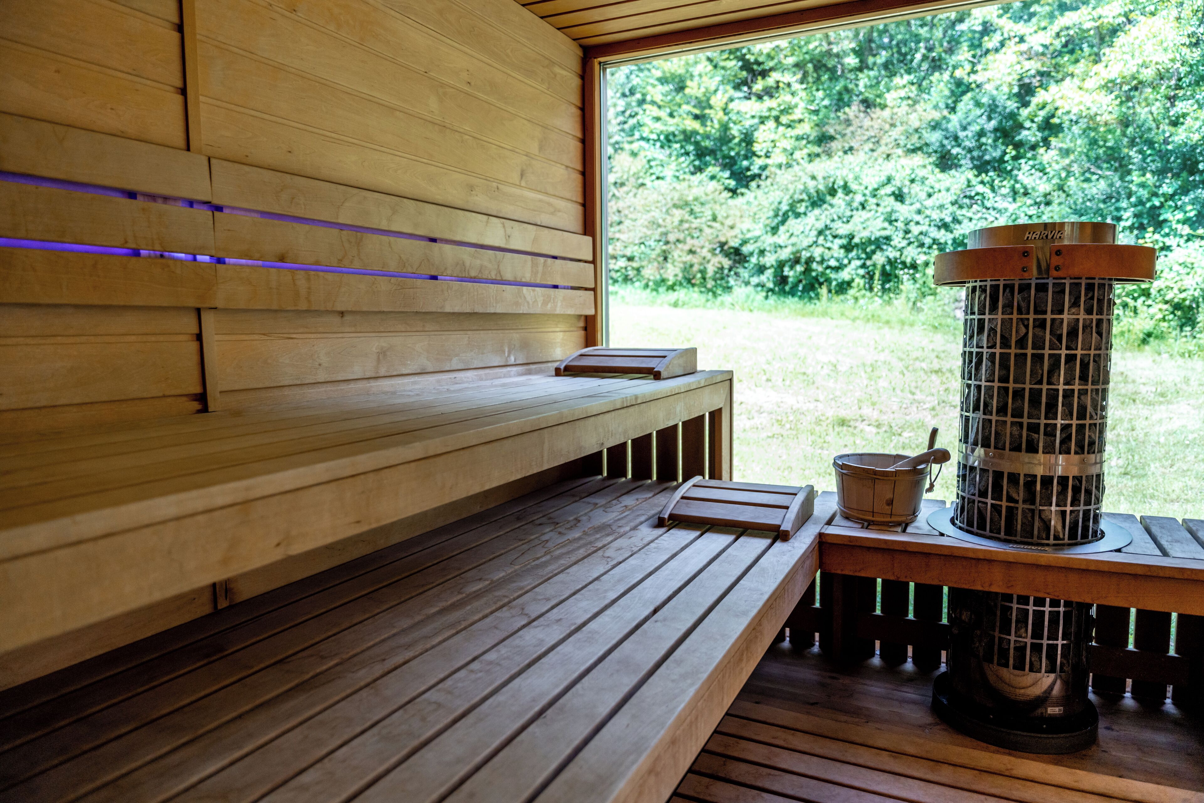 Un sauna en bois, doté de bancs, d’un appareil de chauffage et d’une fenêtre donnant sur la verdure.