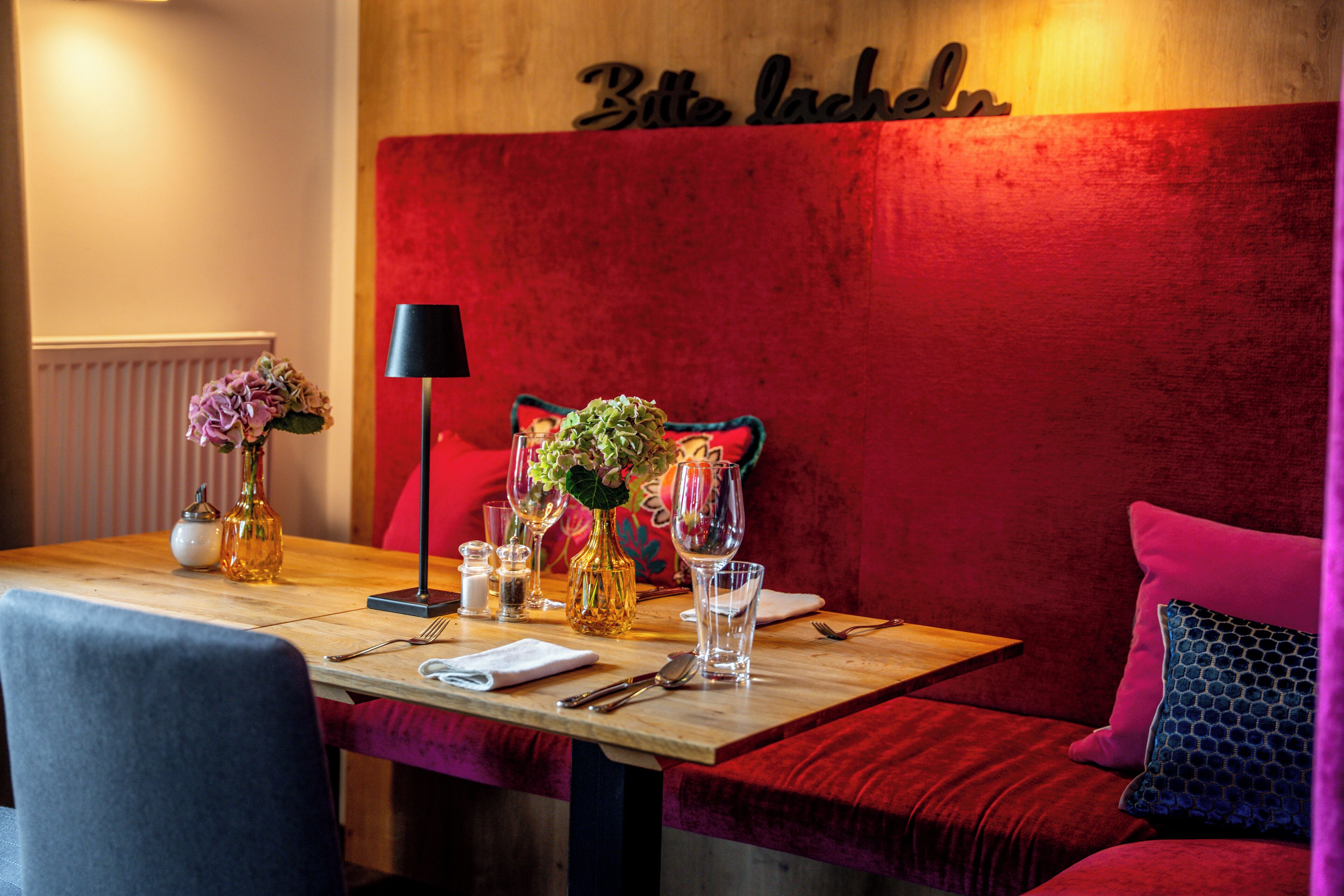 A dining area with a wooden table set for two, a red upholstered bench, and a wall with a sign.