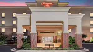 Ingresso di un hotel Hampton Inn con facciata in mattoni, insegna al neon e una panchina di fronte.