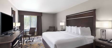 A hotel room with a large bed, a desk with a TV, a chair, a lamp, and a nightstand.