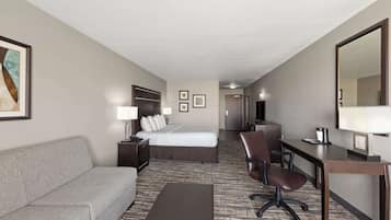 A hotel room with a bed, a sofa, a desk with a chair, a large mirror, and a carpeted floor.