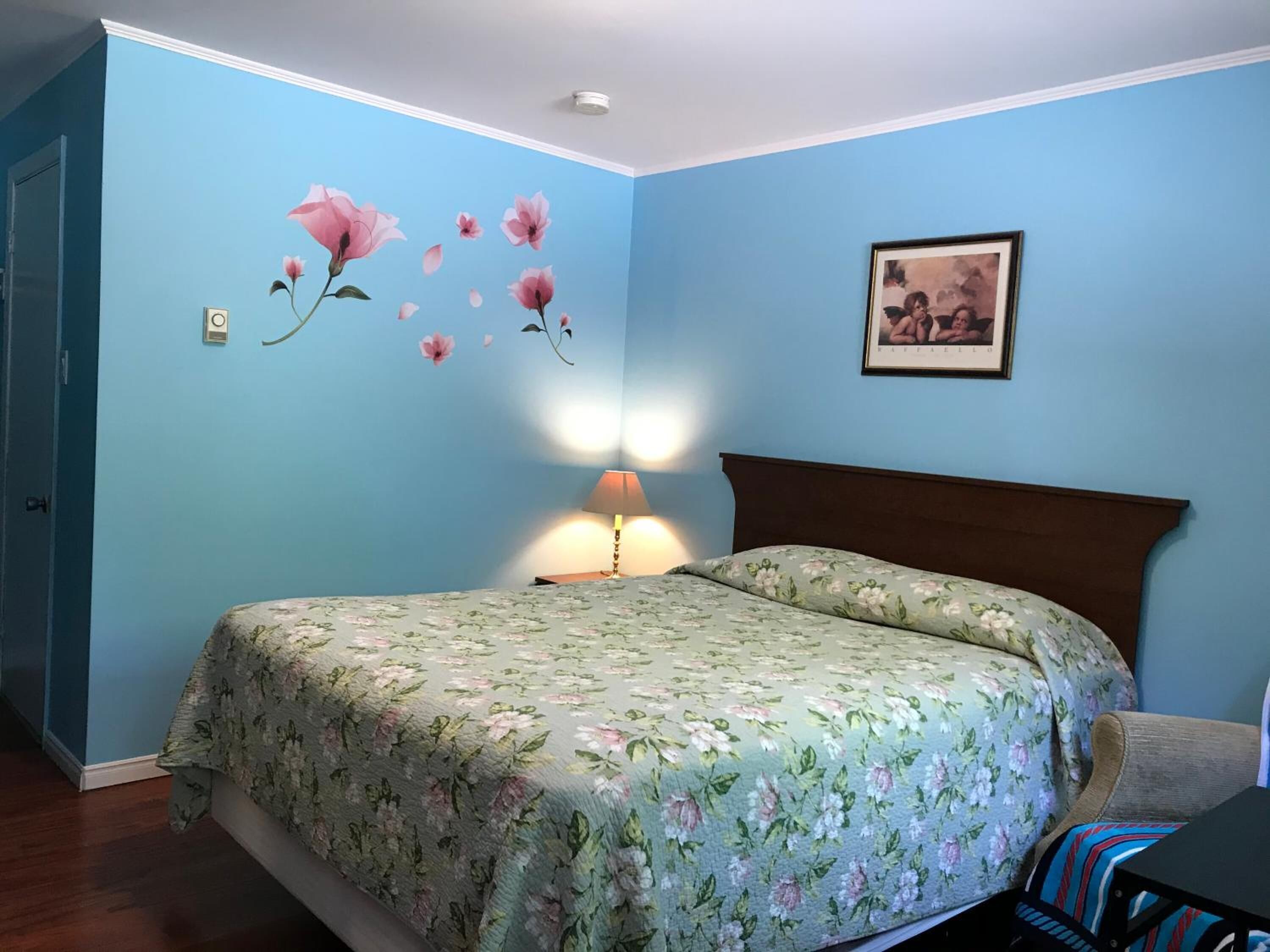 A bedroom with a floral bedspread, a nightstand with a lamp, a framed picture on the wall, and a door to the left.