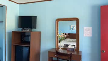 Una habitación con un escritorio de madera, televisor, refrigerador pequeño, espejo y puerta roja.