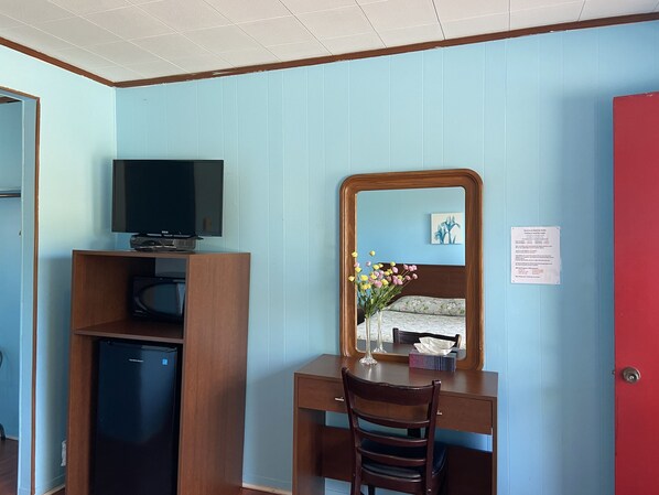 A room with a wooden desk, a television, a small refrigerator, a mirror, and a red door.