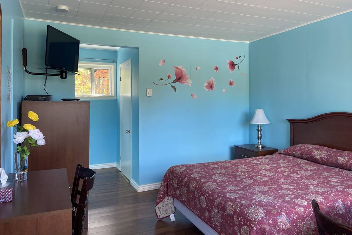 A bedroom with a bed, a desk, a chair, a TV mounted on the wall, and a window.