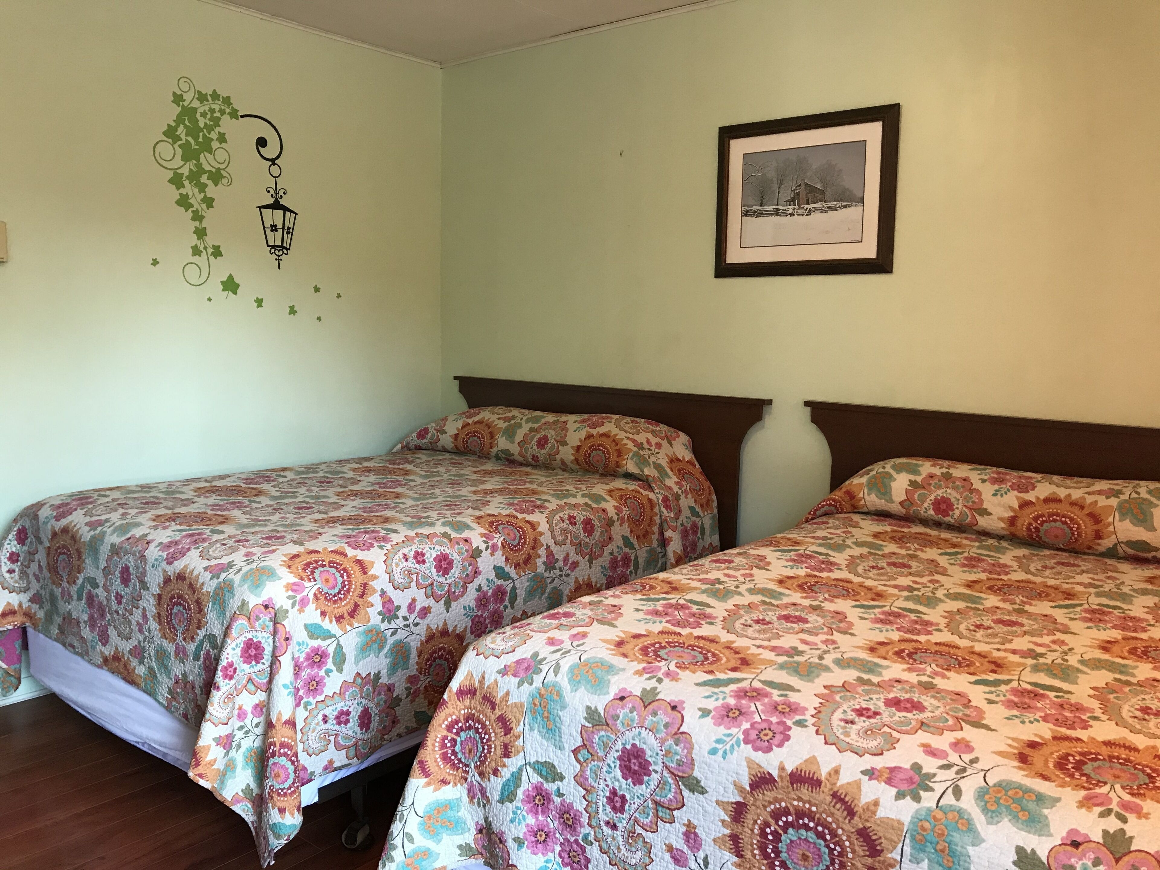 A room with two beds, each with floral-patterned bedding, a framed picture on the wall, and a wall decal of a hanging lamp and ivy.