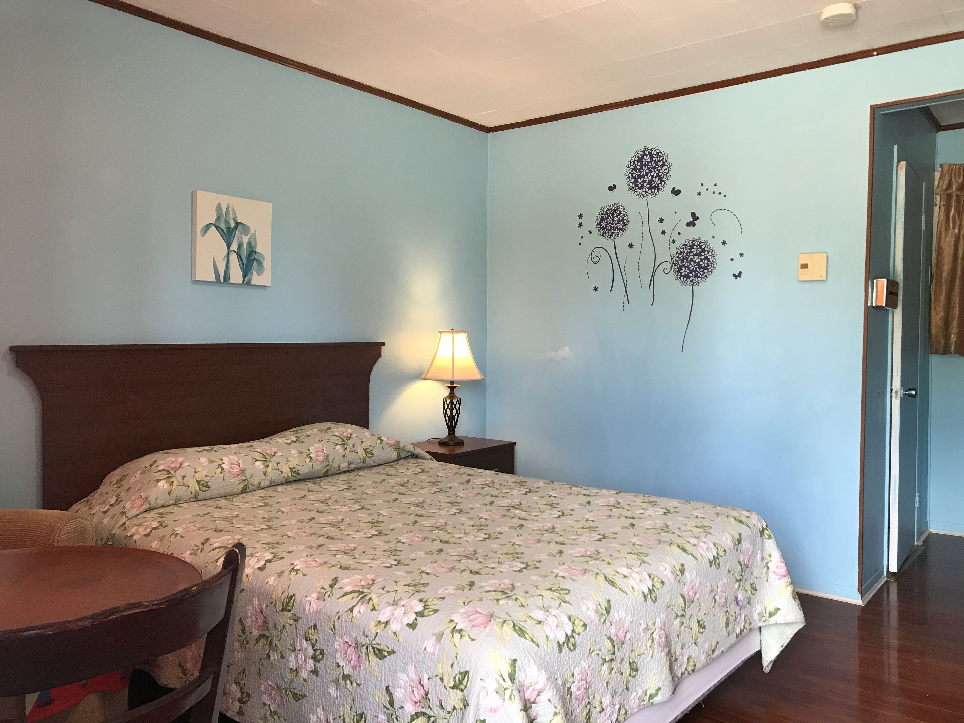 Un dormitorio con edredón estampado, cabecera de madera, lámpara de noche, una mesita con una silla y un adorno en la pared.