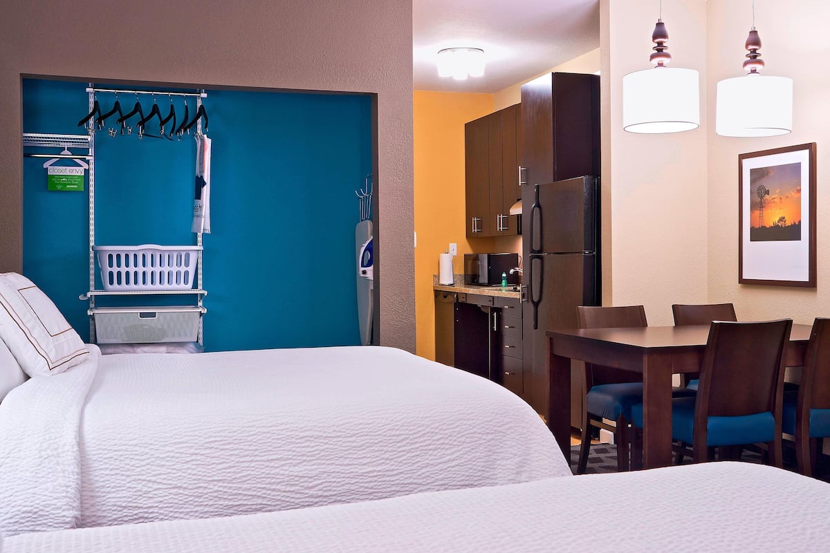 A hotel room with a bed, a dining area with chairs and a table, a kitchen area with a refrigerator, and a blue wall with a closet and hanging items.
