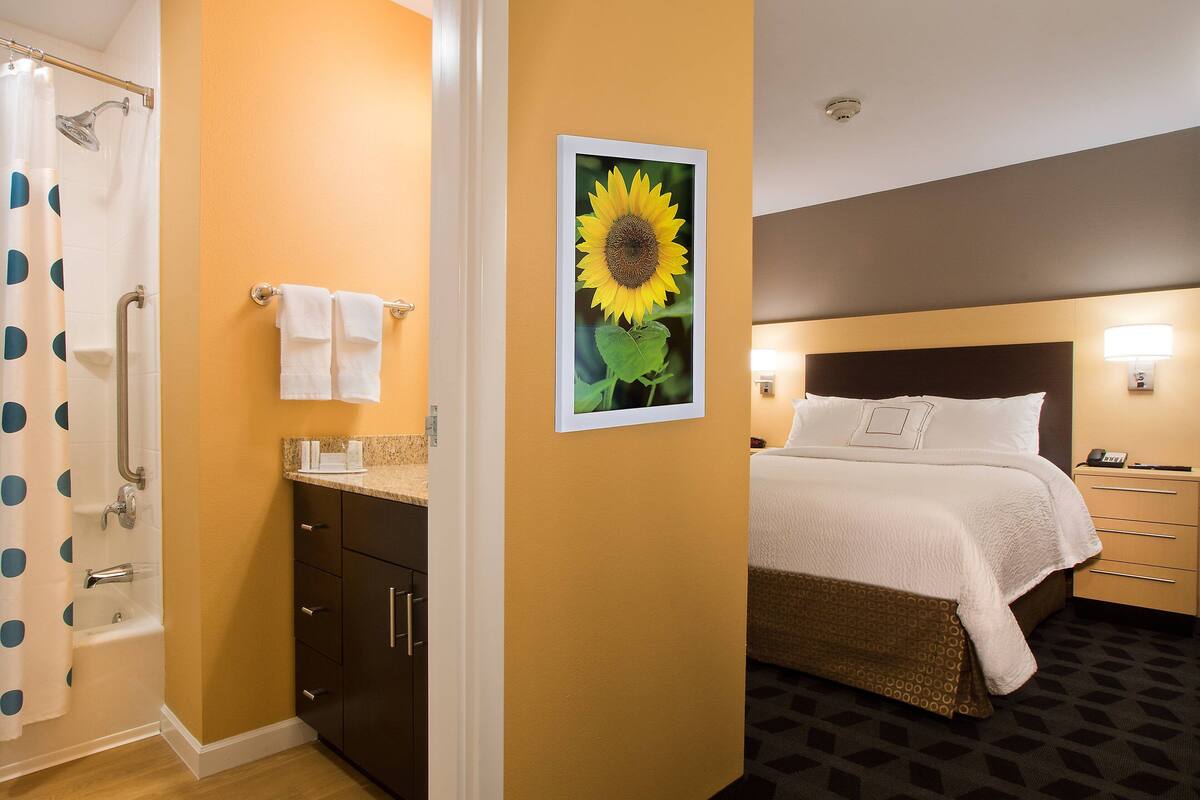 A hotel room with a shower, a sink, a bed, and a wall-mounted picture of a sunflower.