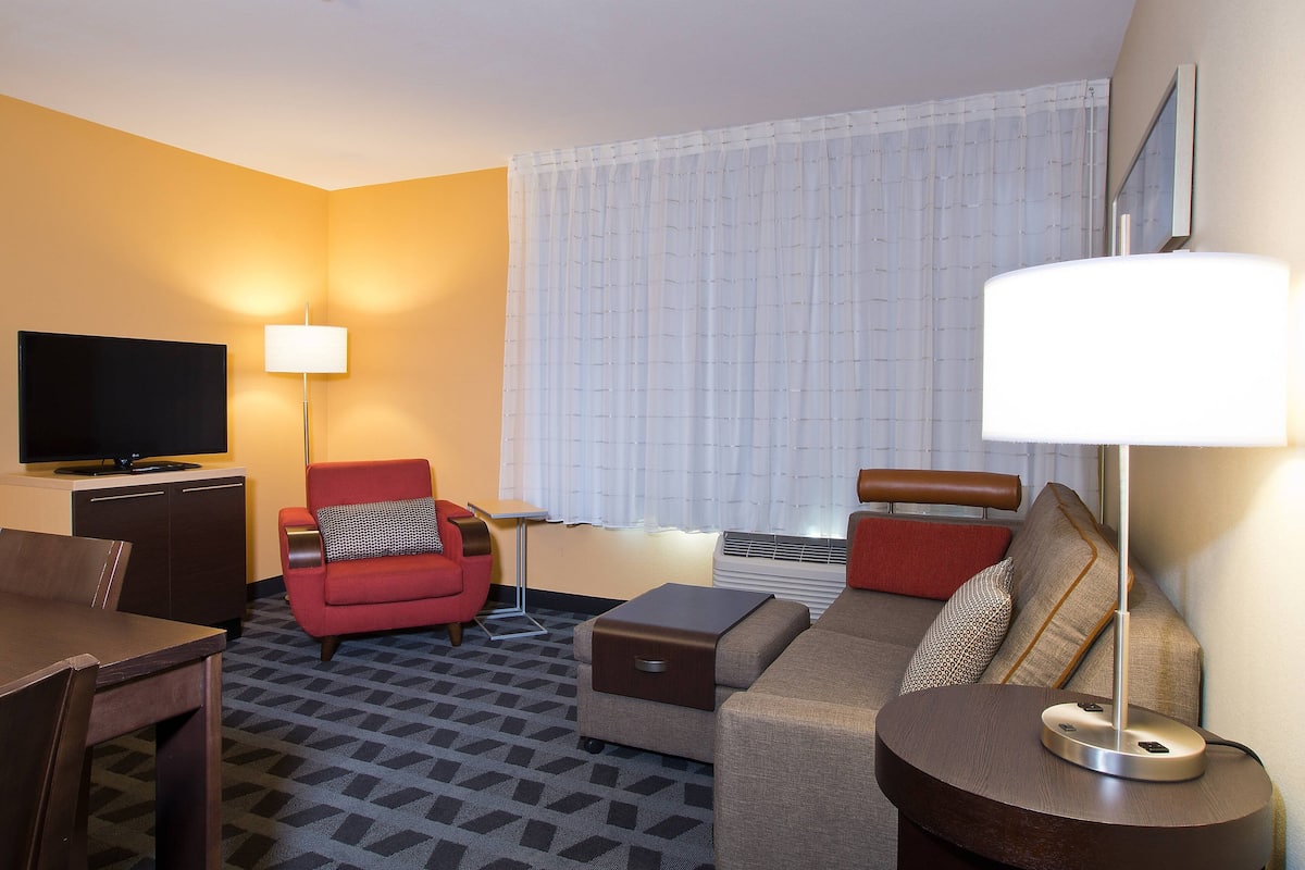 A hotel room with a TV, red armchair, sofa, and a lamp.