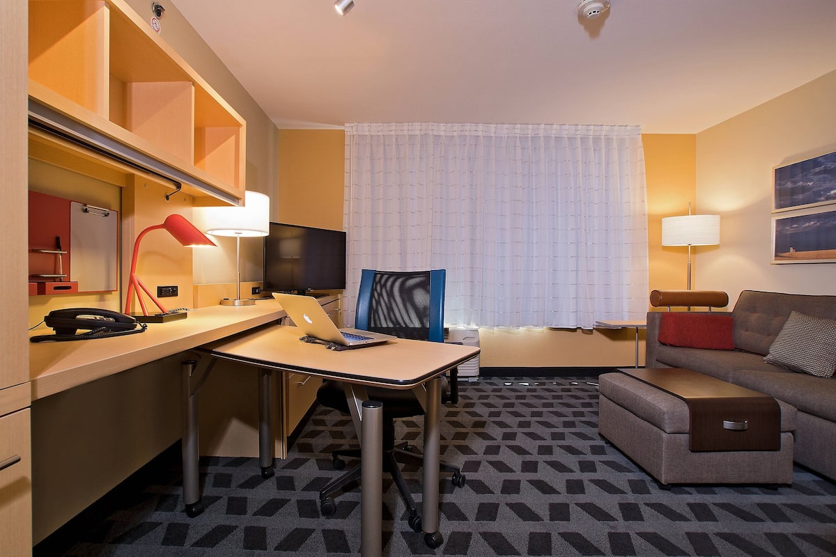 A hotel room with a desk, chair, sofa, and a large window with curtains.