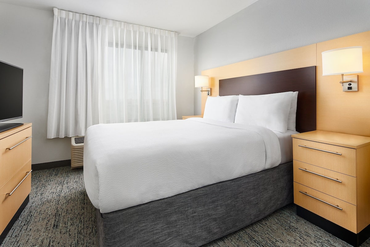 A hotel room with a large bed, a bedside table with a lamp, a TV, and a window with curtains.