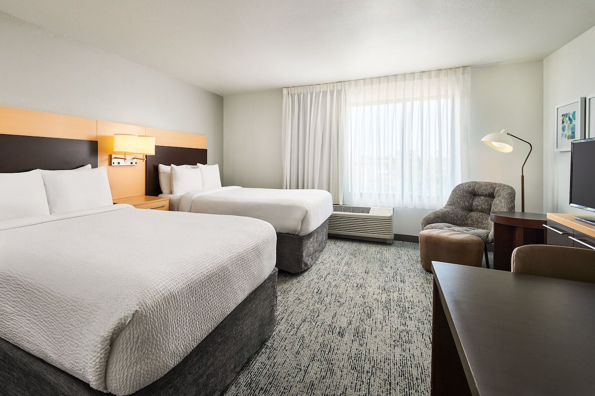 A hotel room with two beds, a desk, a chair, a lamp, and a large window with curtains.