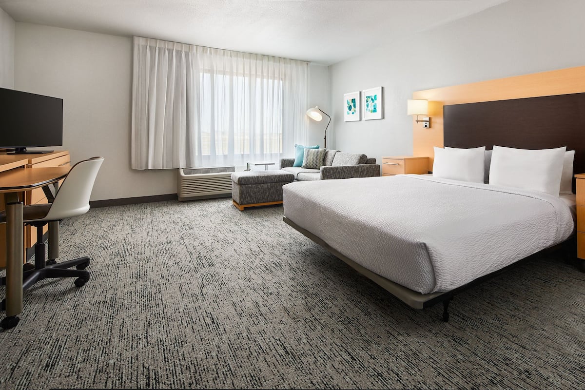 A hotel room with a bed, a desk with a chair, a TV, a lamp, and a window with curtains.