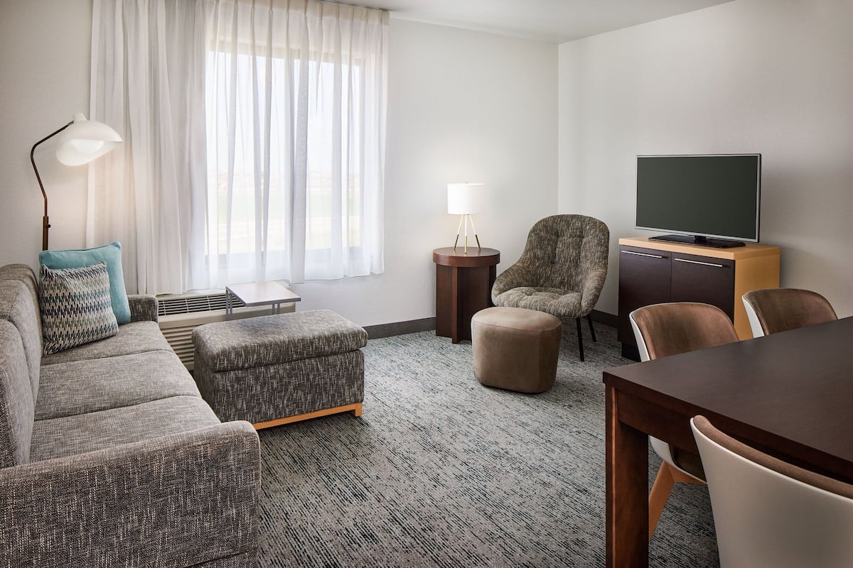 A modern hotel room with a sofa, ottoman, armchair, and a flat-screen TV on a wooden stand.