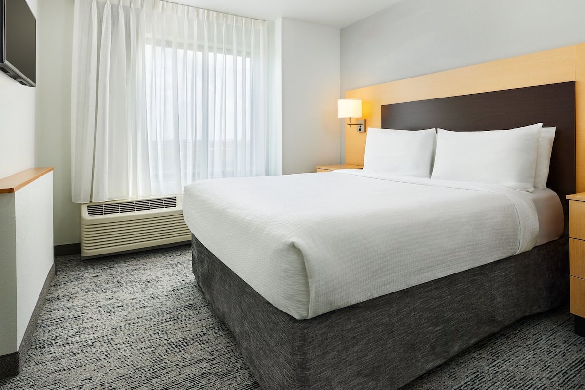 A hotel room with a large bed, a bedside table, a lamp, a window with curtains, and a wall-mounted TV.