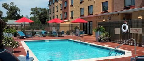 Una piscina con lettini e ombrelloni rossi, circondata da una recinzione e un edificio con numerose finestre.