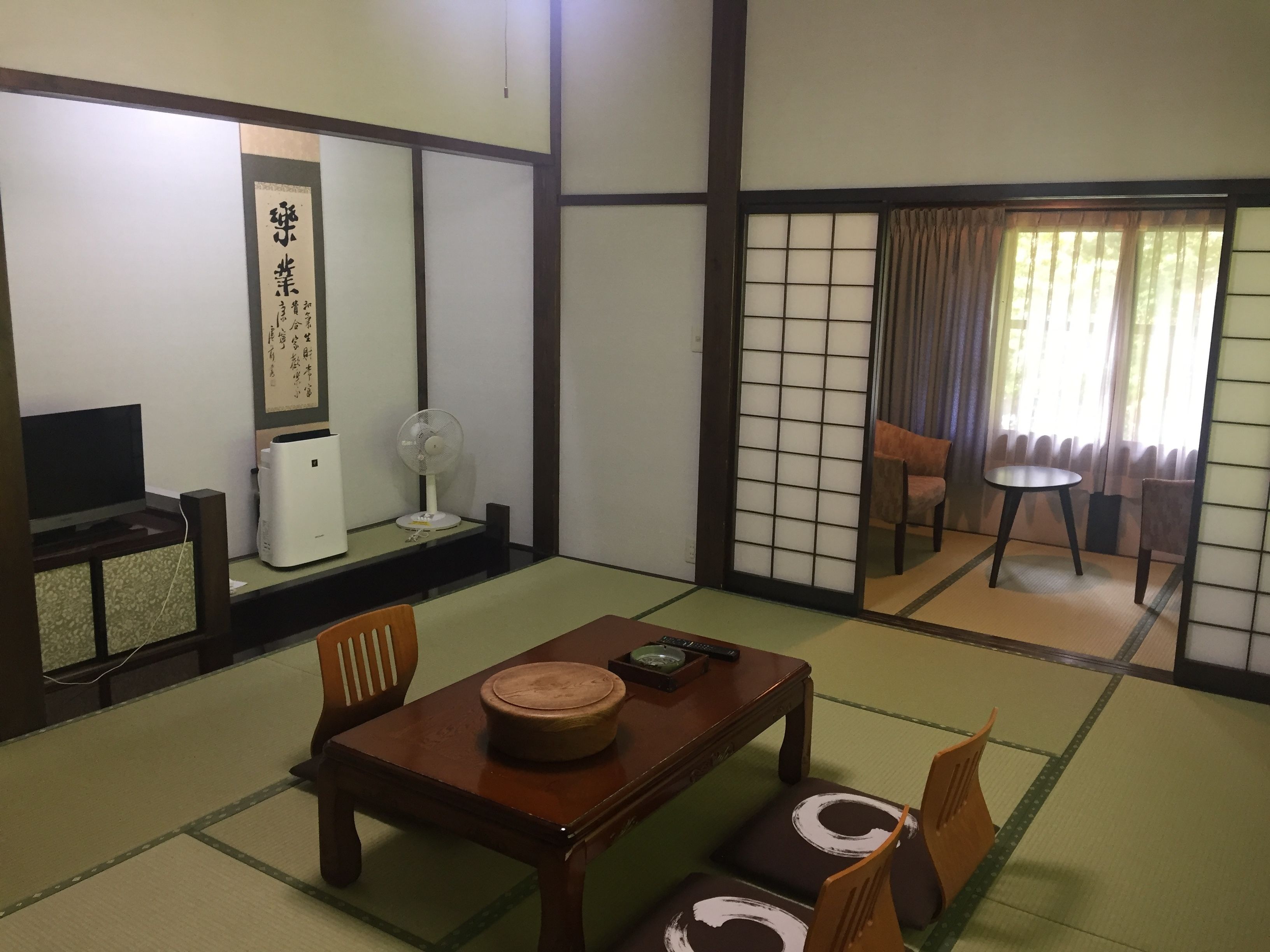 A traditional Japanese room with tatami flooring, a low table with cushions, a television, a fan, and a sliding door with a view of the outside.