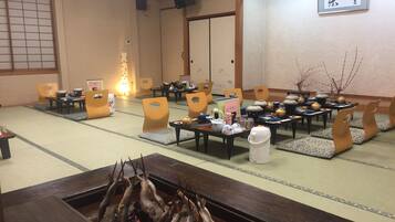 A traditional Japanese room with tatami flooring, low tables, and chairs. There is a fire pit with fish, a projector screen, and a sign with Japanese characters.