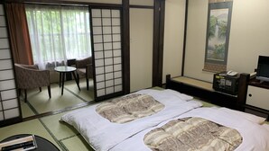 Ett traditionellt japanskt rum med tatamimattor på golvet, en futonmadrass, ett litet bord med en fjärrkontroll, en stol och ett skrivbord med en telefon och en tv.