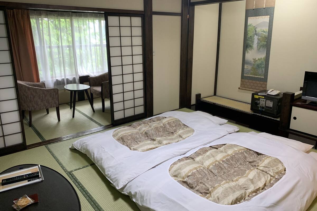 A traditional Japanese-style room with tatami flooring, a futon bed, a small table with a remote control, a chair, and a desk with a phone and a TV.