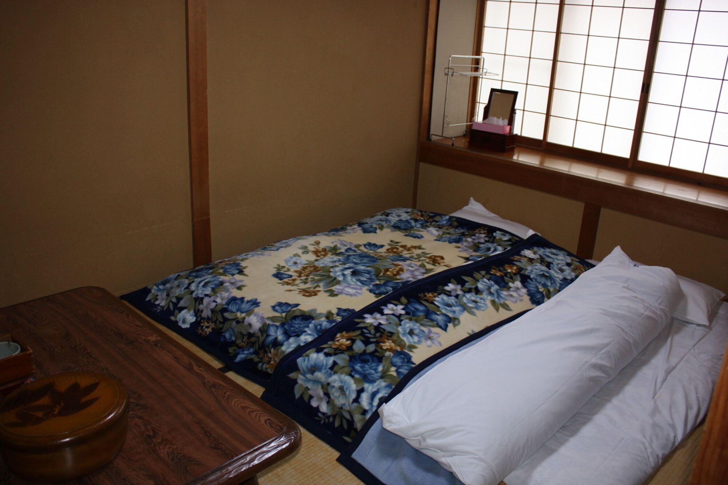 Quarto pequeno e bem arrumado, com cama, mesa de madeira e janela com vista.