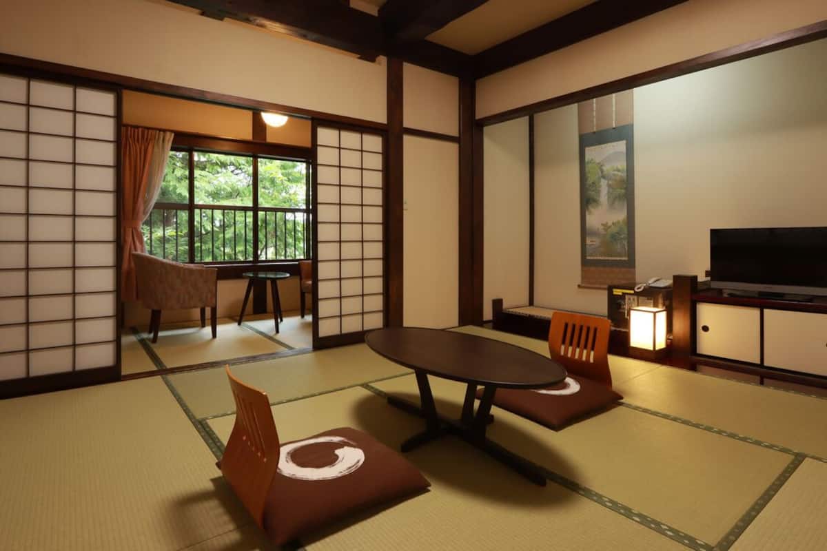 A traditional Japanese room with tatami flooring, a low table, chairs, a television, and a sliding door leading to a balcony.