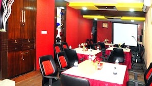A conference room with red walls, wooden cabinets, and black chairs with red cushions. Tables are set with water bottles and floral centerpieces. - Hotel Rose Garden (Dhaka)