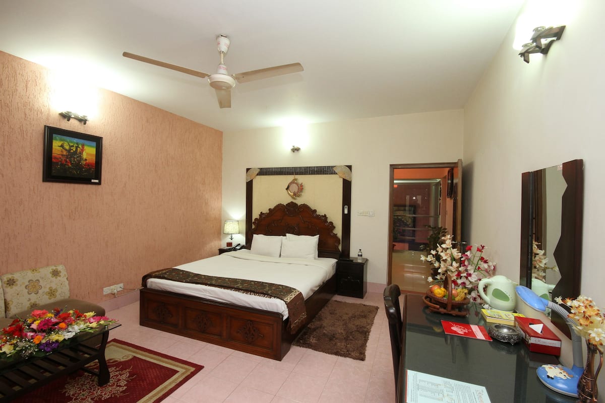 A bedroom with a bed, a desk, a chair, a ceiling fan, a nightstand, a lamp, a mirror, a vase with flowers, and a framed picture on the wall.