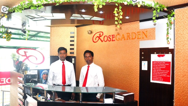 A reception desk at Rosegarden with two staff members, a glass partition, and a sign listing services like room booking and ticket confirmation. - Hotel Rose Garden (Dhaka)