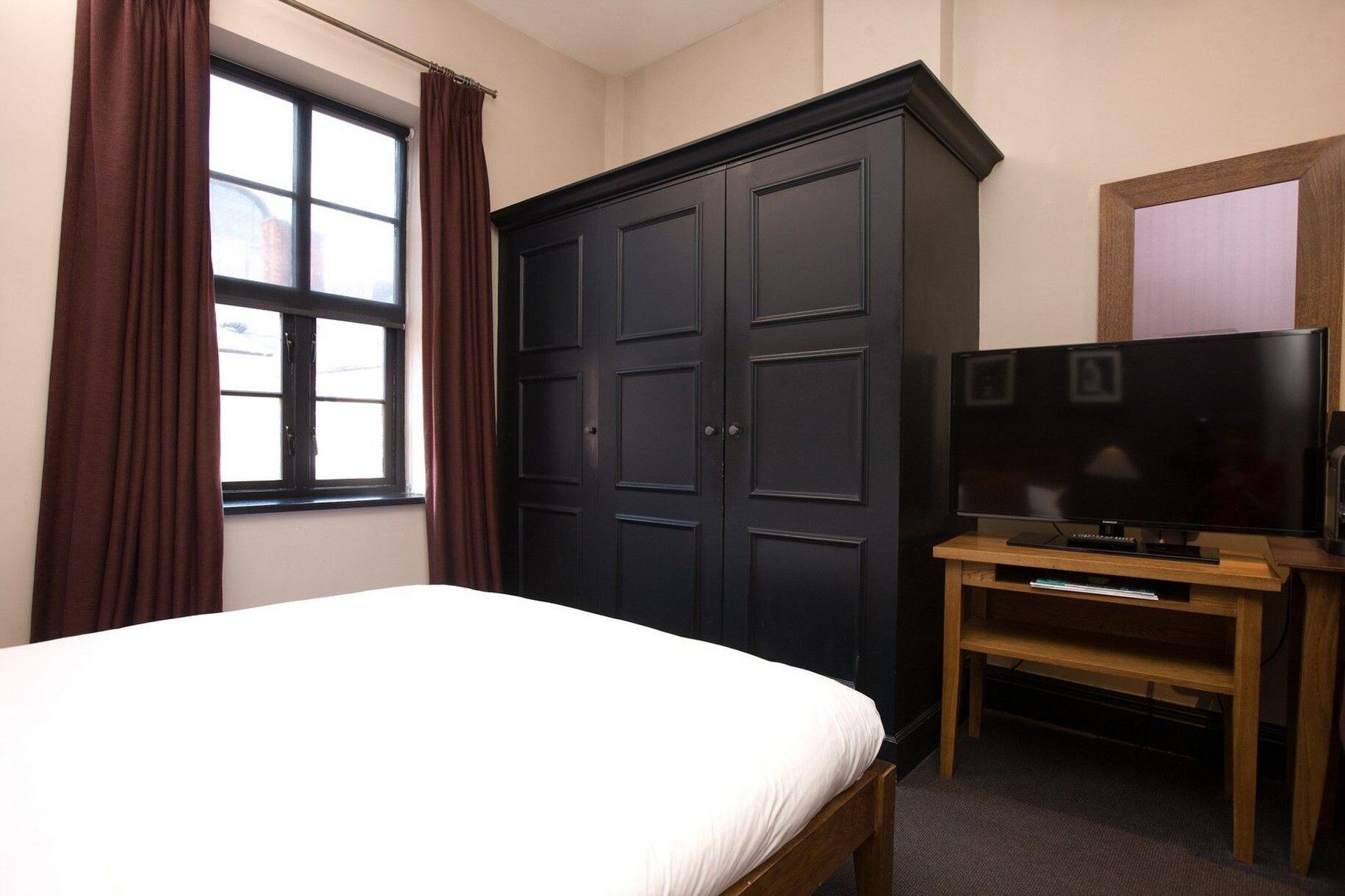 Una habitación de hotel con cama, armario, televisor sobre un soporte de madera y ventana con cortinas.