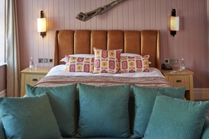 Uma cama com cabeceira marrom, almofadas azul-turquesa e roupa de cama estampada. Há duas mesas de cabeceira com abajures e um telefone.