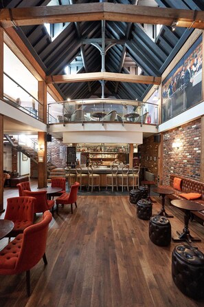 A spacious indoor area with a high ceiling, wooden beams, and a bar in the center. There are multiple seating areas with chairs and tables, and a staircase leading to an upper level. - Hotel du Vin Brighton (Brighton)