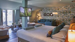 Ein modernes Hotelzimmer mit einem großen Bett, einer Sitzecke inklusive Sofa, einem Flachbildfernseher, einer Badewanne und einem großen Fenster.