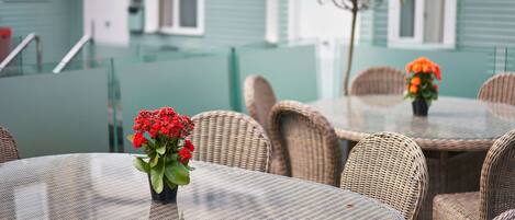 A dining area with wicker chairs and a glass table, a small potted plant, and a building with a green facade in the background.
