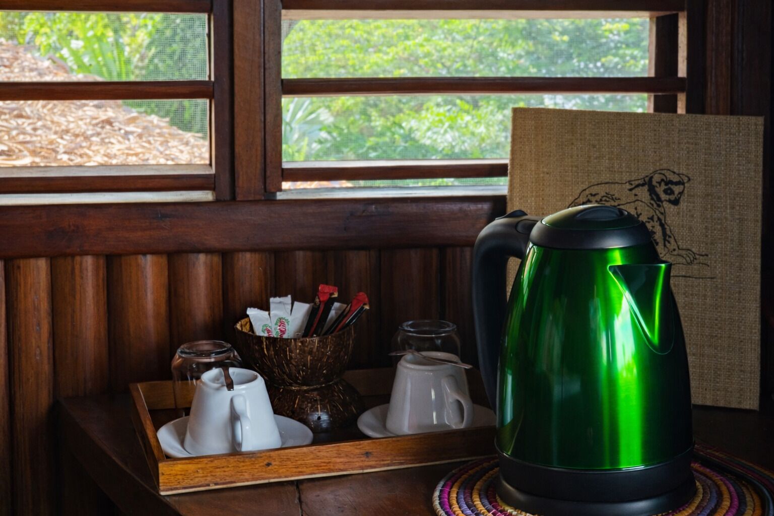 A wooden bench with a tray holding white teapots and a green kettle, a woven basket with utensils, and a window with a view of greenery.