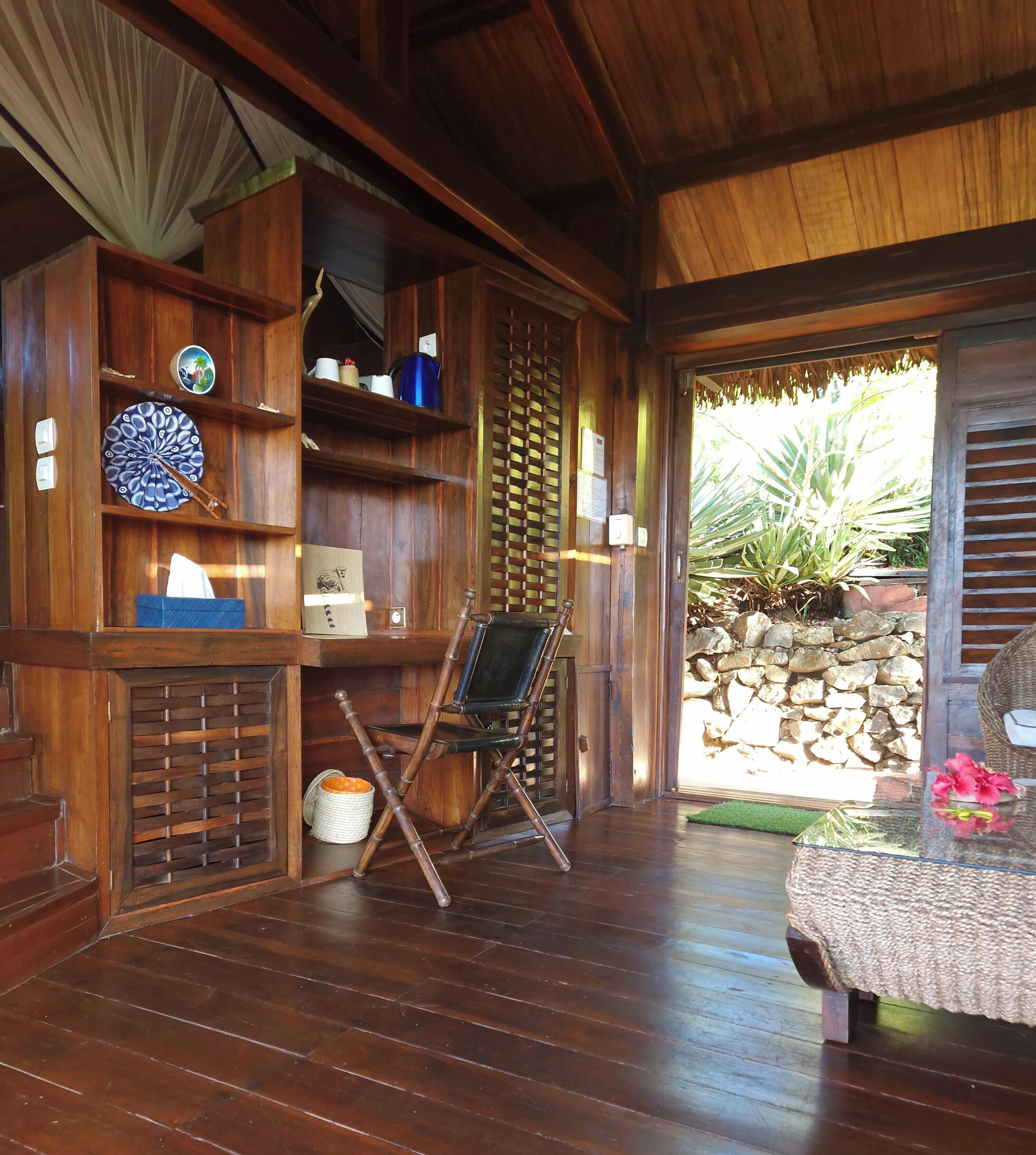 A wooden interior with a wooden floor, a chair, a table with a vase, and a view of the outside.