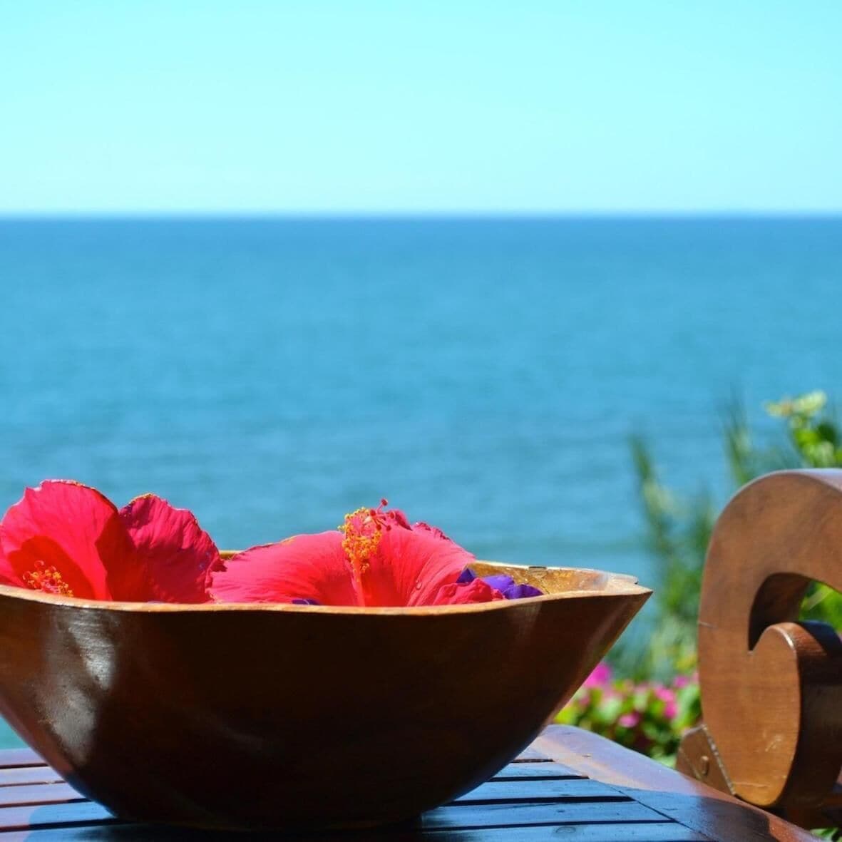 Un tazón de madera con flores rojas sobre una mesa, con vista al océano.