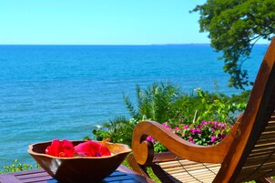Un banco de madera con asiento tejido y un cuenco de flores rojas sobre una mesa de madera junto al mar.