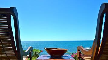 Two wooden lounge chairs facing a clear blue sea, with a bowl on a small table between them.