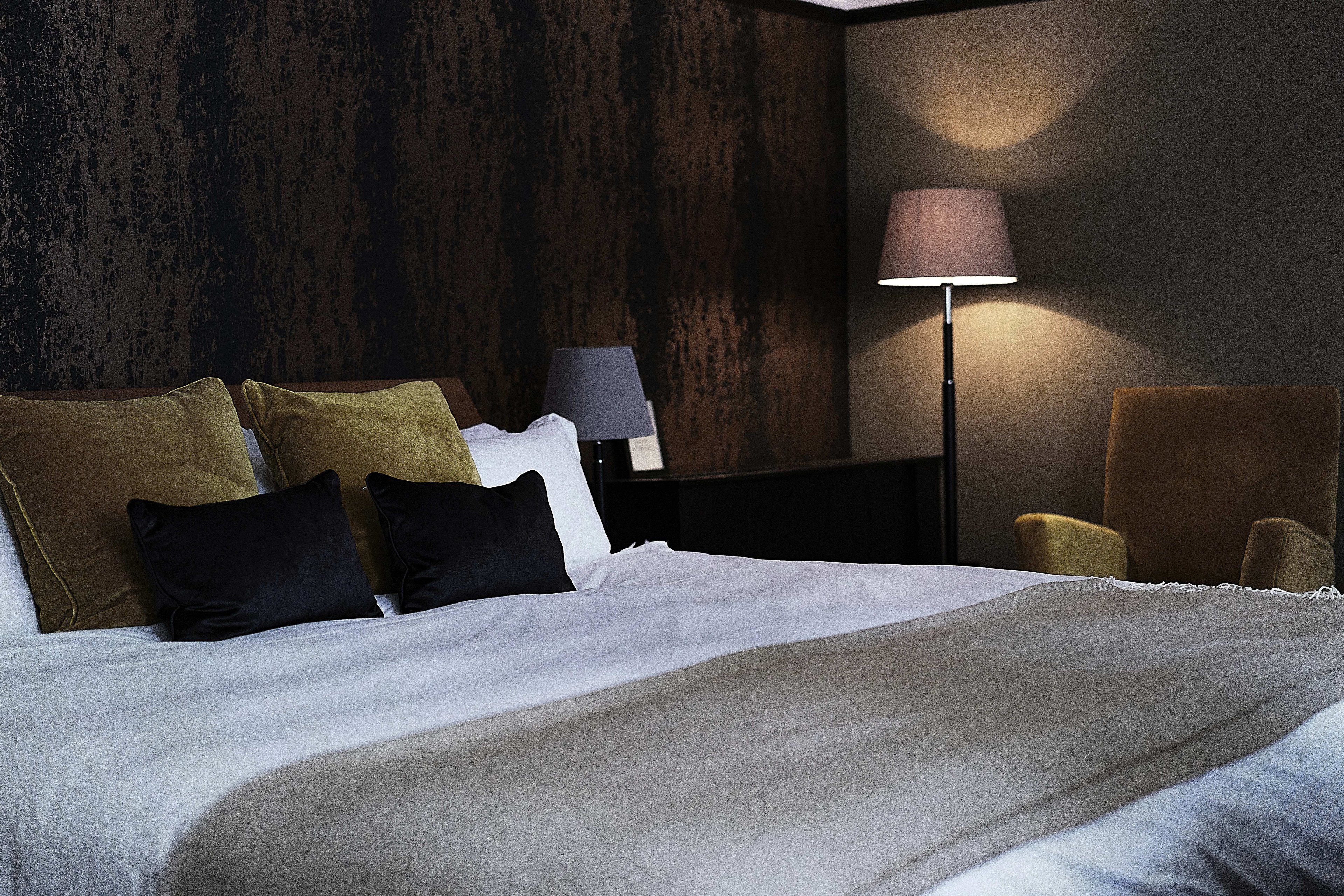 A hotel room with a bed, pillows, a nightstand, a lamp, and a chair.