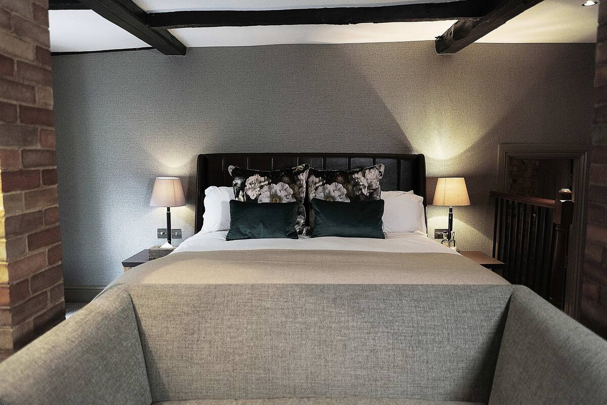 A hotel room with a large bed, two bedside tables with lamps, a brick wall, and a wooden ceiling beam.