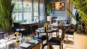 A well-lit dining area with multiple tables set for guests, large windows allowing natural light, and potted plants adding greenery. - Hotel du Vin & Bistro Cambridge (Cambridge)