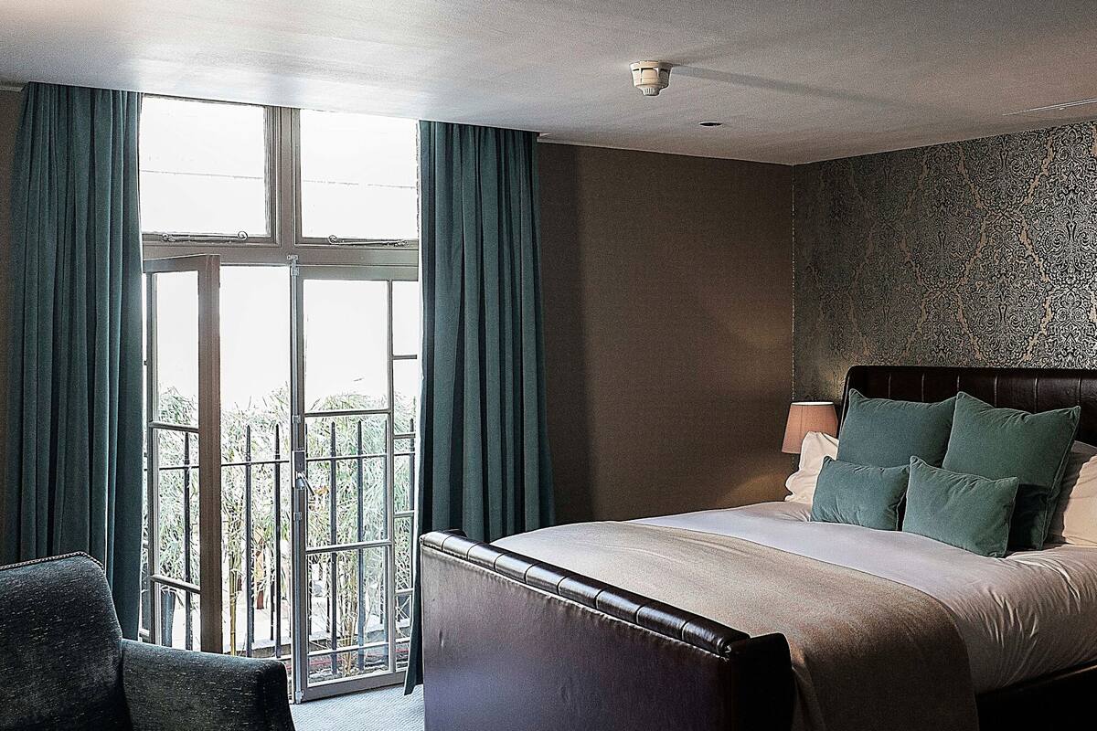 A hotel room with a large bed, a chair, a window with curtains, and a patterned wall.