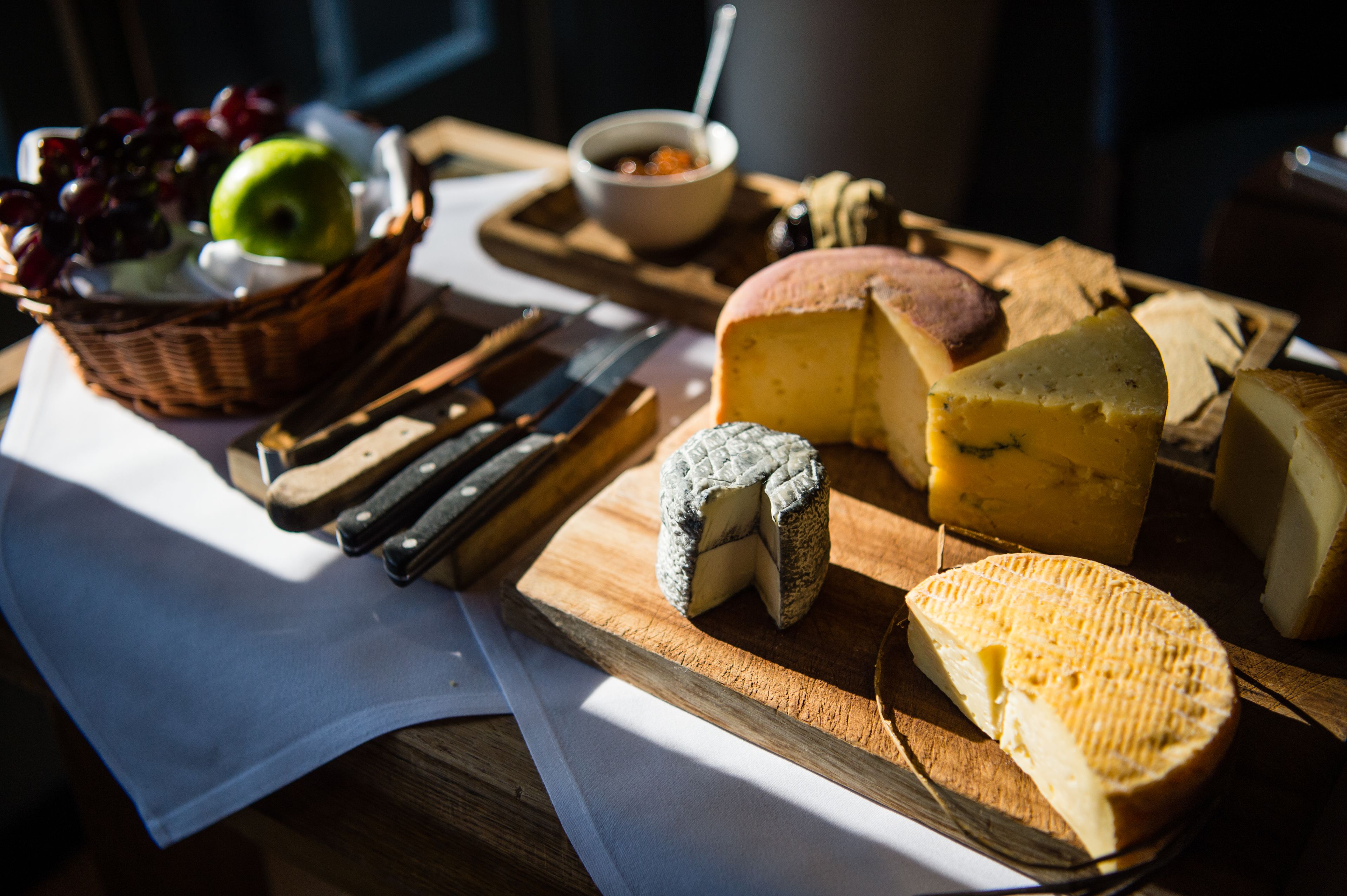 A variety of cheeses and fruits on a wooden board with knives, suggesting a cheese tasting event.